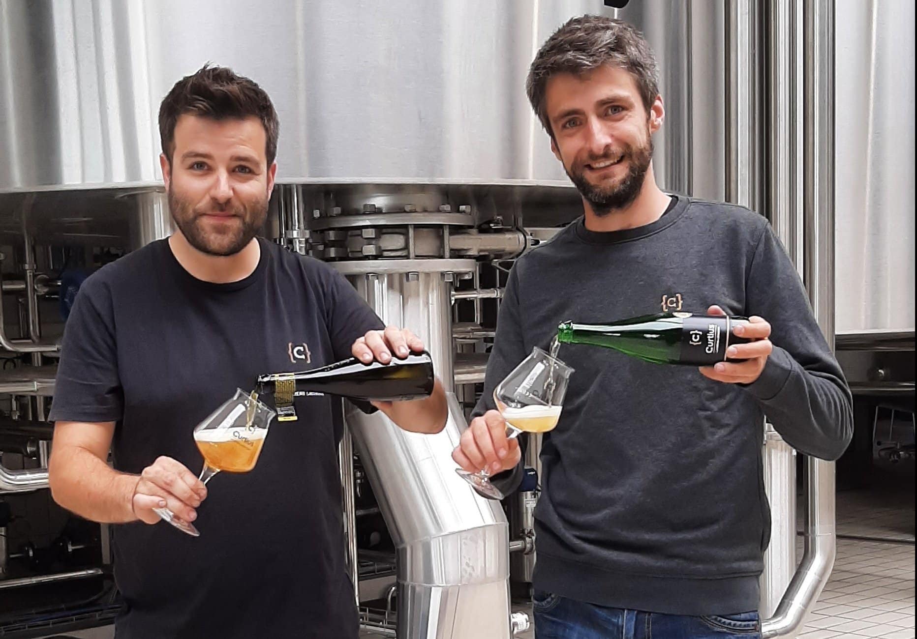 Deux hommes en t-shirt versent de la bière depuis des bouteilles dans des verres devant des cuves de brasserie