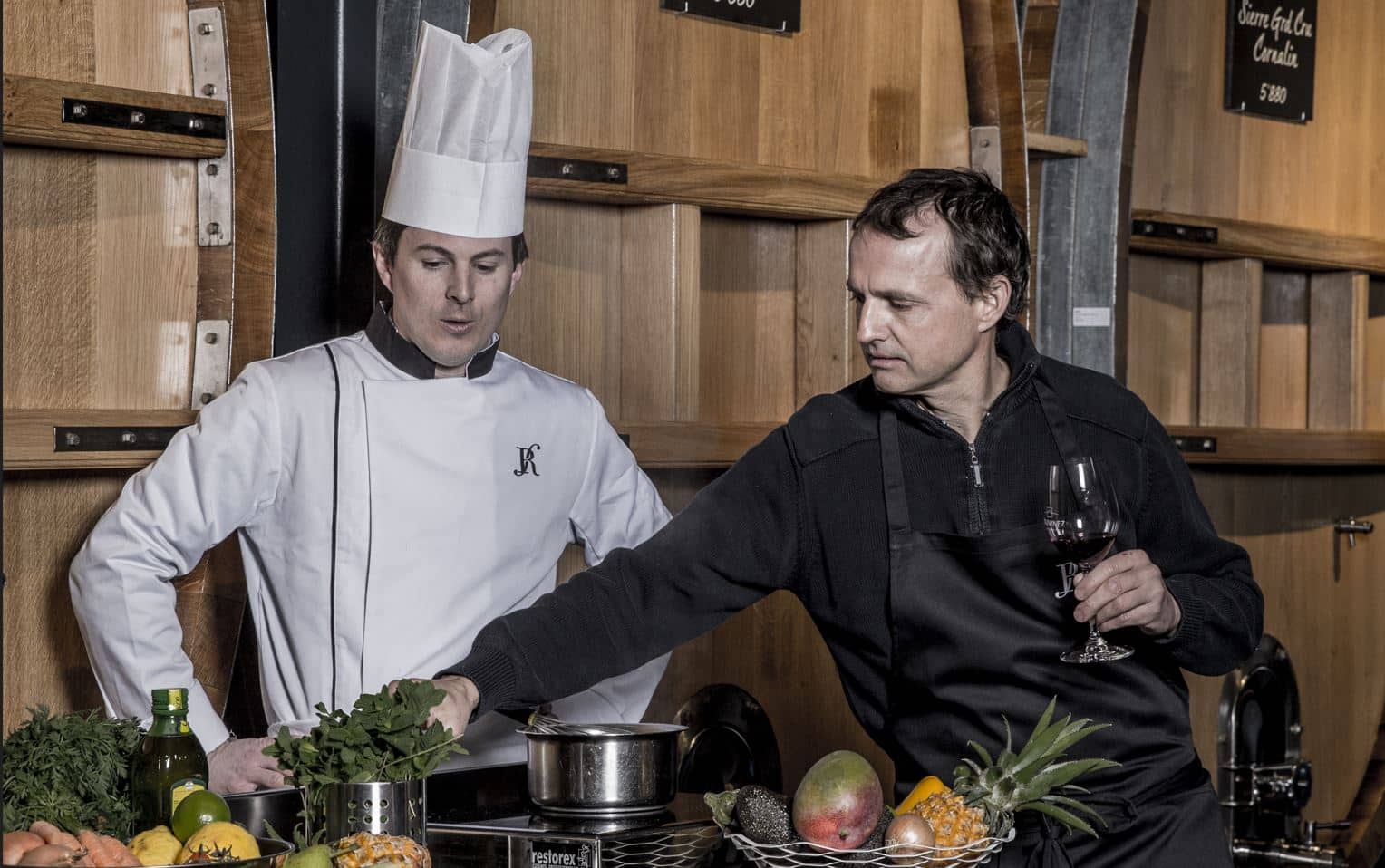 Un chef en toque et un homme en veste noire dégustant un verre de vin rouge préparent un plat dans une cave viticole