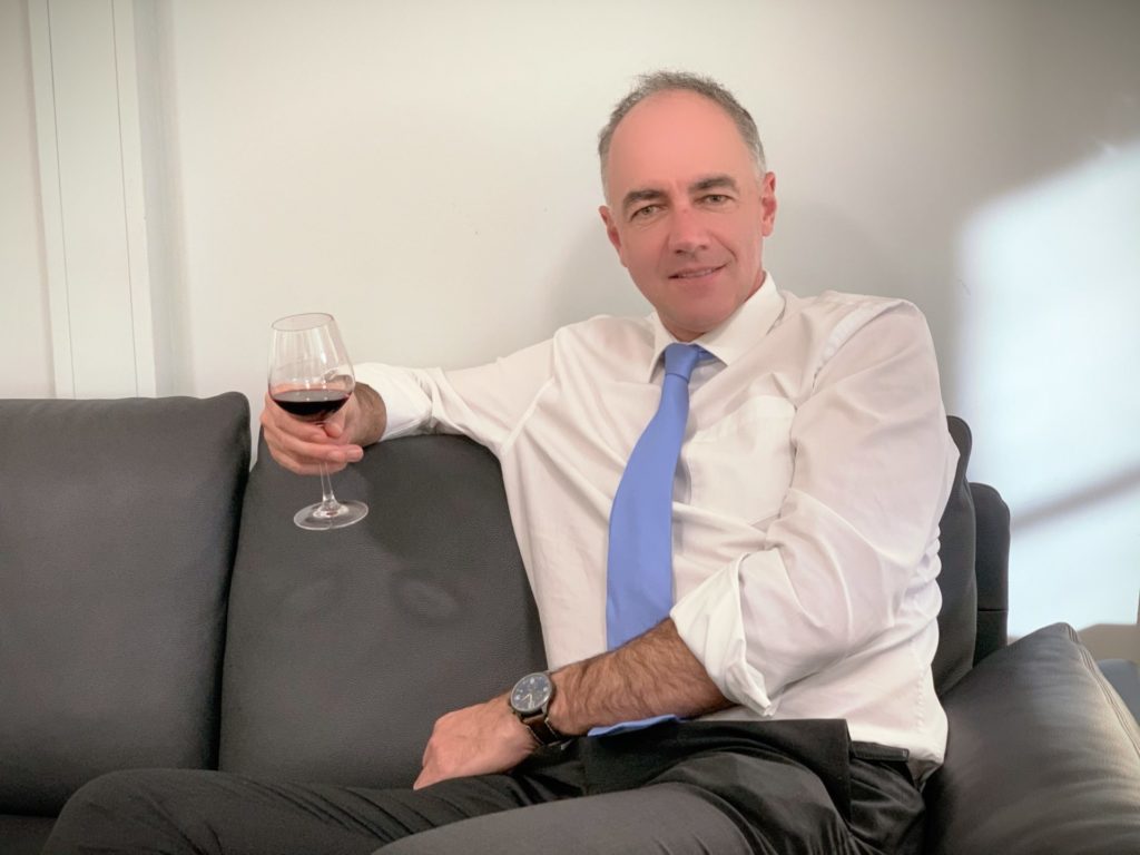 Mann in weißem Hemd und blauer Krawatte, der ein Glas Rotwein hält und auf einem Sofa sitzt