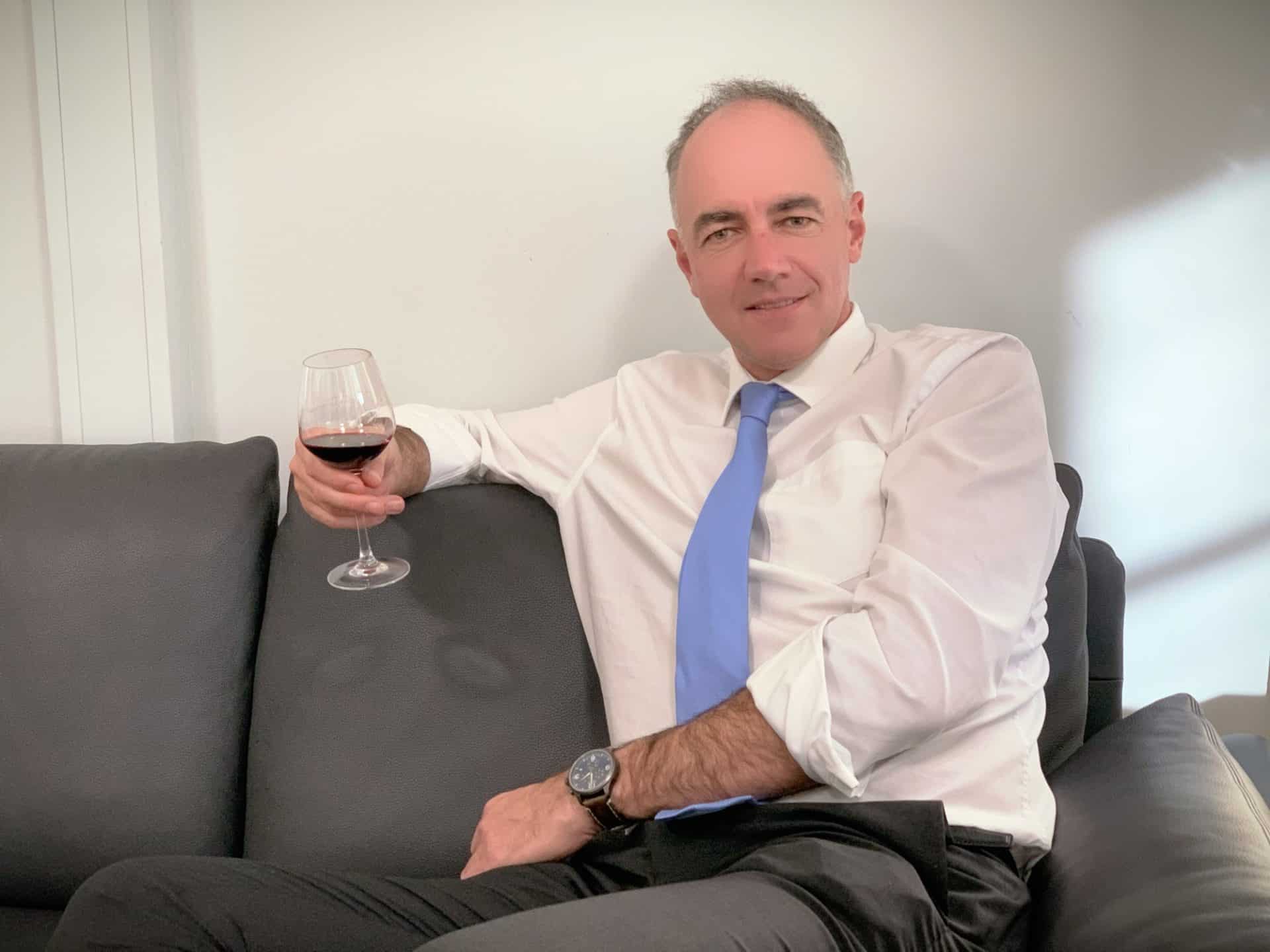 Homme en chemise blanche et cravate bleue tenant un verre de vin rouge assis sur un canapé